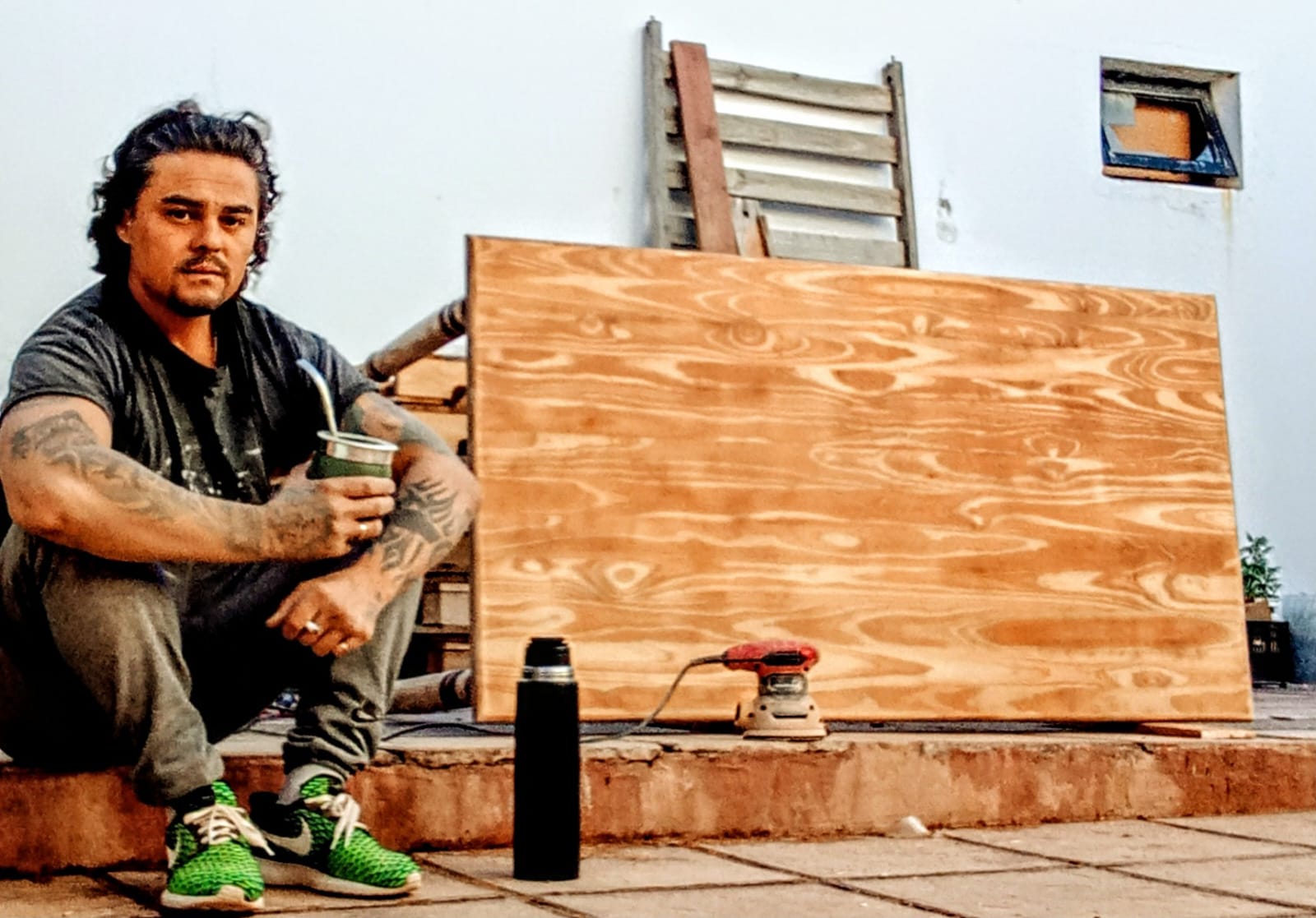 Mauricio tomando mate junto a una tabla lijada, descansando en el taller