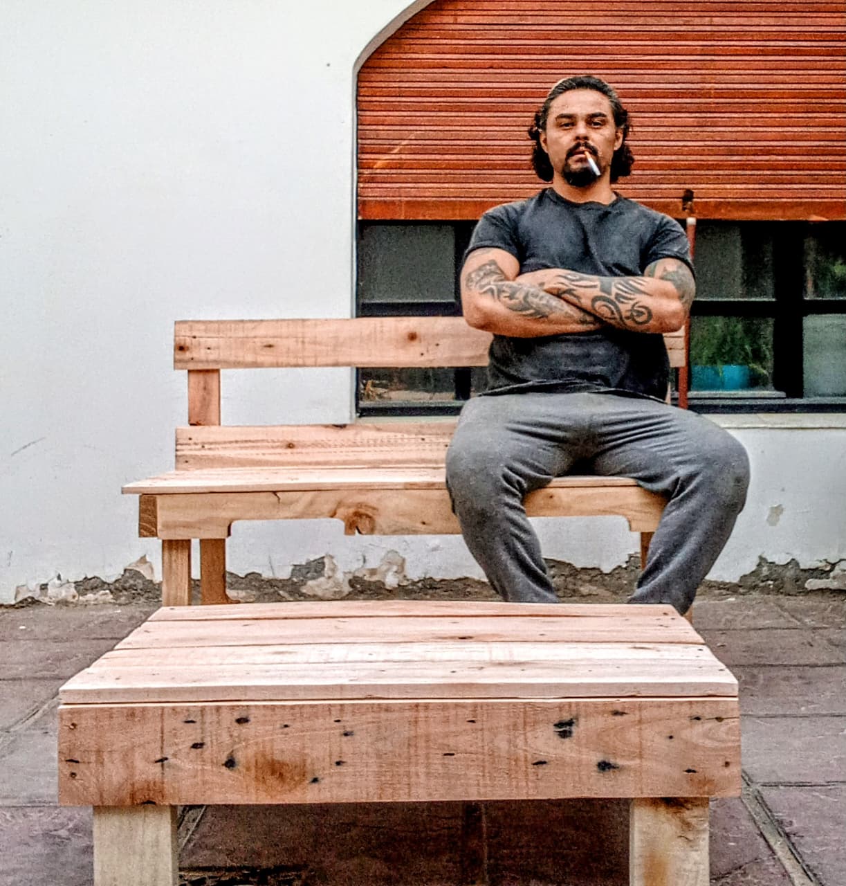 Mauricio sentado en un banco de pallet junto a una mesa ratona hecha a mano en su patio
